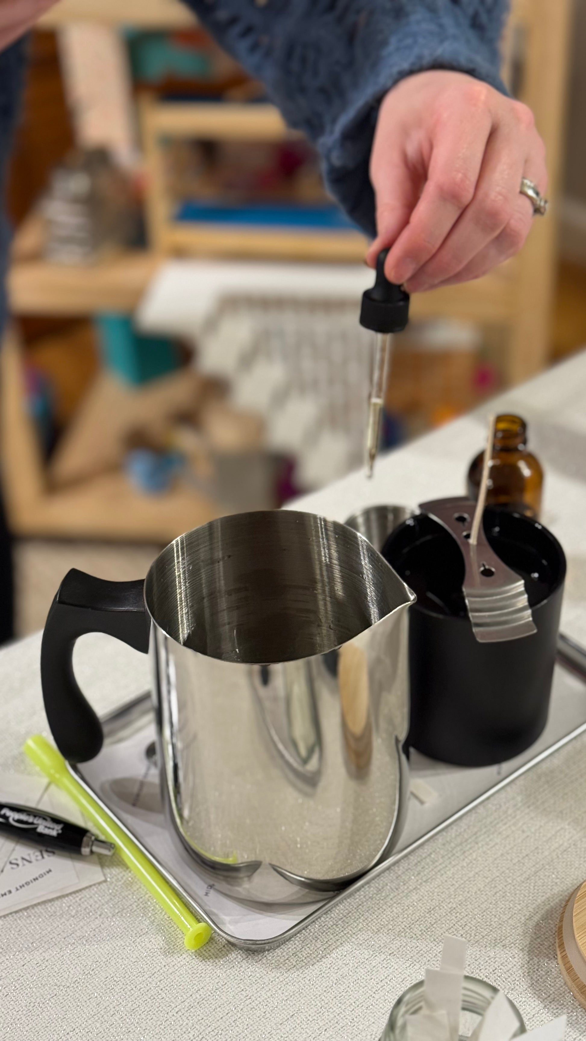 A person holding a dropper filled with liquid over a stainless steel container on a silver tray, with various other items arranged around it, indicating a candle making activity.