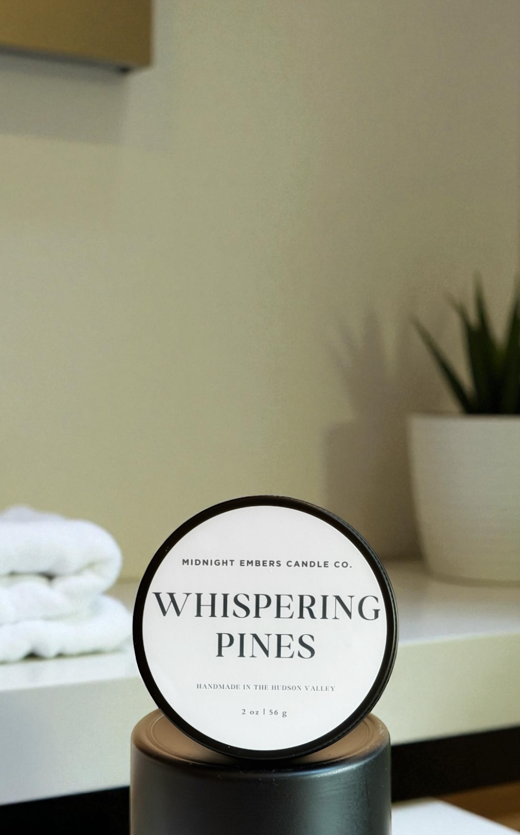 Candle labeled 'Whispering Pines' on a stand with a blurred background of a room.