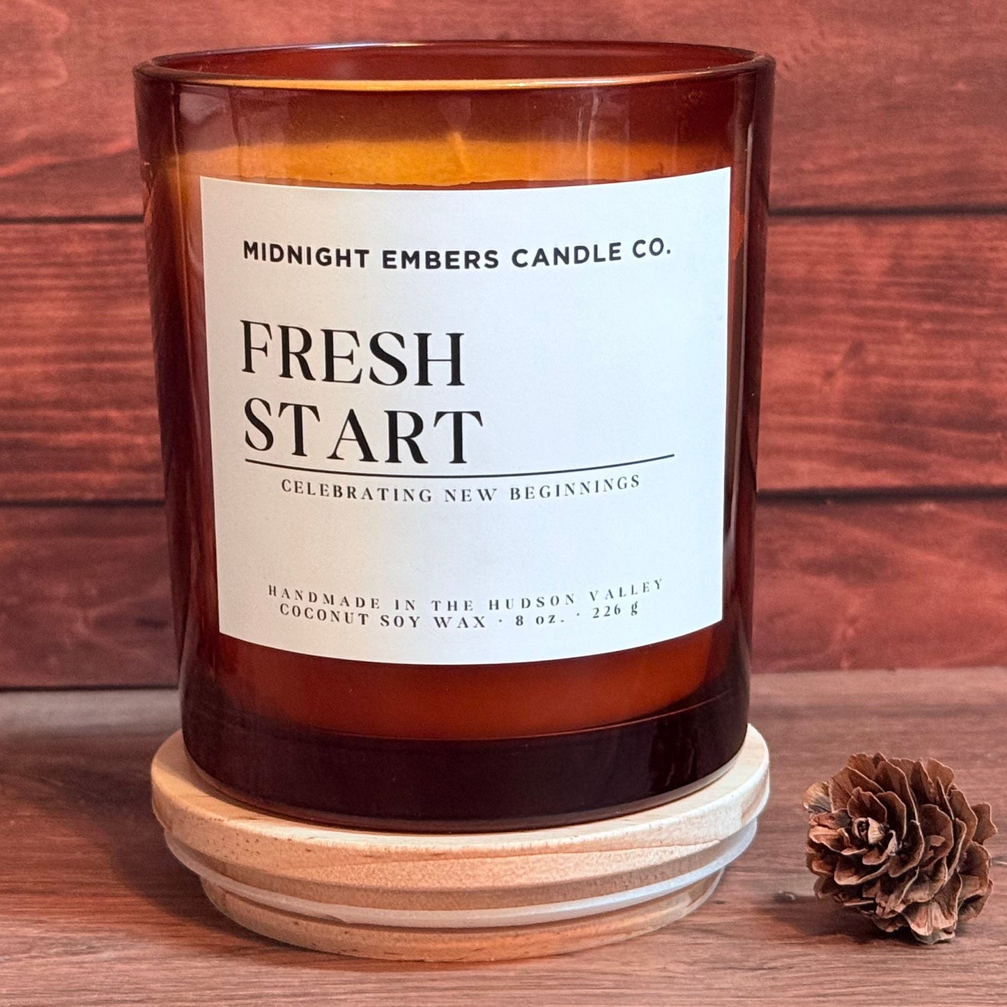 An amber glass jar candle with a white label reading 'MIDNIGHT EMBERS CANDLE CO. FRESH START' sits on a round wooden coaster against a rustic wood plank background, with a small pinecone beside it.