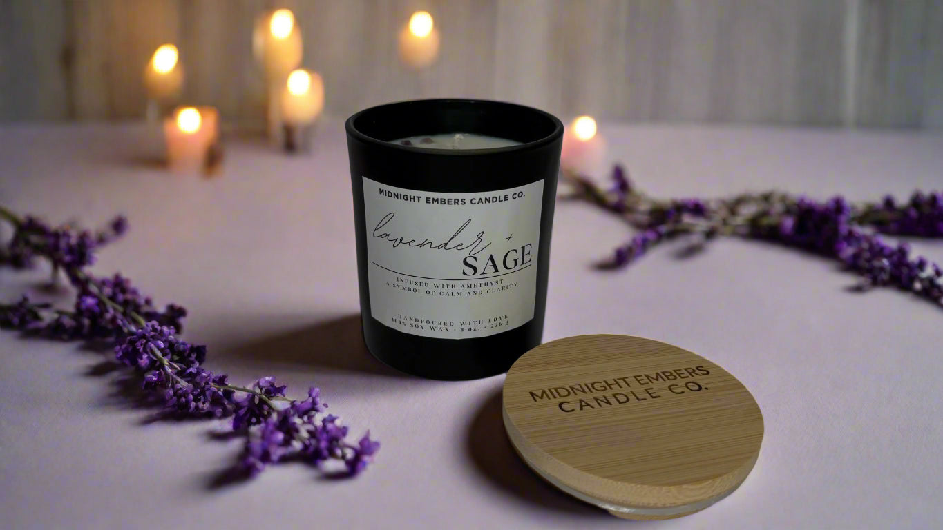 A matte black jar candle with a bamboo wooden lid, labeled 'Lavender + Sage'. It is surrounded by scattered lavender petals and other candles in the background, suggesting a relaxing, aromatic environment.