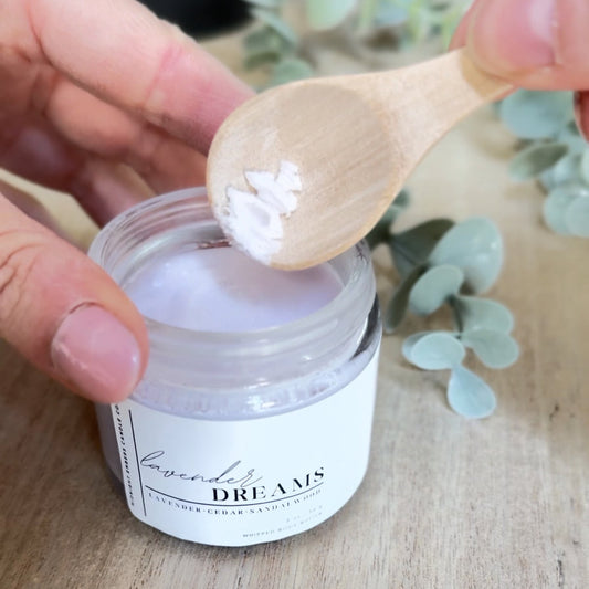 Person using a wooden spoon to scoop out a white powder from a jar labeled 'Dreams' on a wooden surface with eucalyptus leaves.
