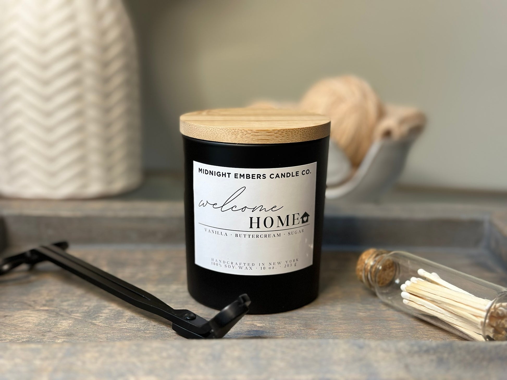 A matte black glass jar candle with a wooden lid labeled 'Welcome Home', alongside a wick trimmer and a small glass bottle with matches.