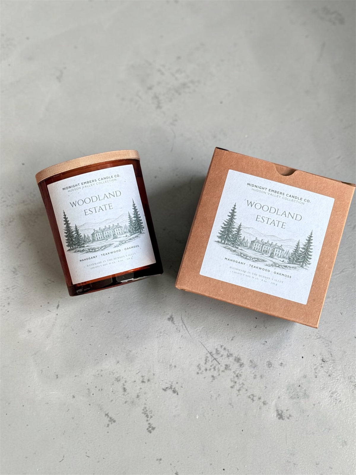 A glass jar candle with a copper-toned lid sits beside its kraft paper box on a light gray textured surface. Both pieces display a white label with a black line drawing of pine trees and the text 'Woodland Estate' and 'Midnight Forest Candle Co.