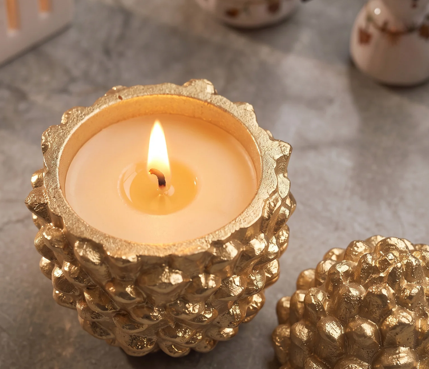 A lit cream-colored candle in a textured metallic gold holder, shown on a gray stone-like surface with a matching gold decorative object partially visible in the foreground.