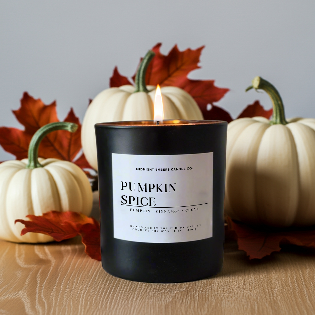 A black candle jar labeled 'Pumpkin Spice' by Midnight Smoker Candle Co, lit and placed on a wooden surface with white pumpkins and red autumn leaves in the background.