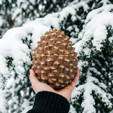 A hand holding a pinecone-shaped container with a metallic gold finish, set against a snowy evergreen background.