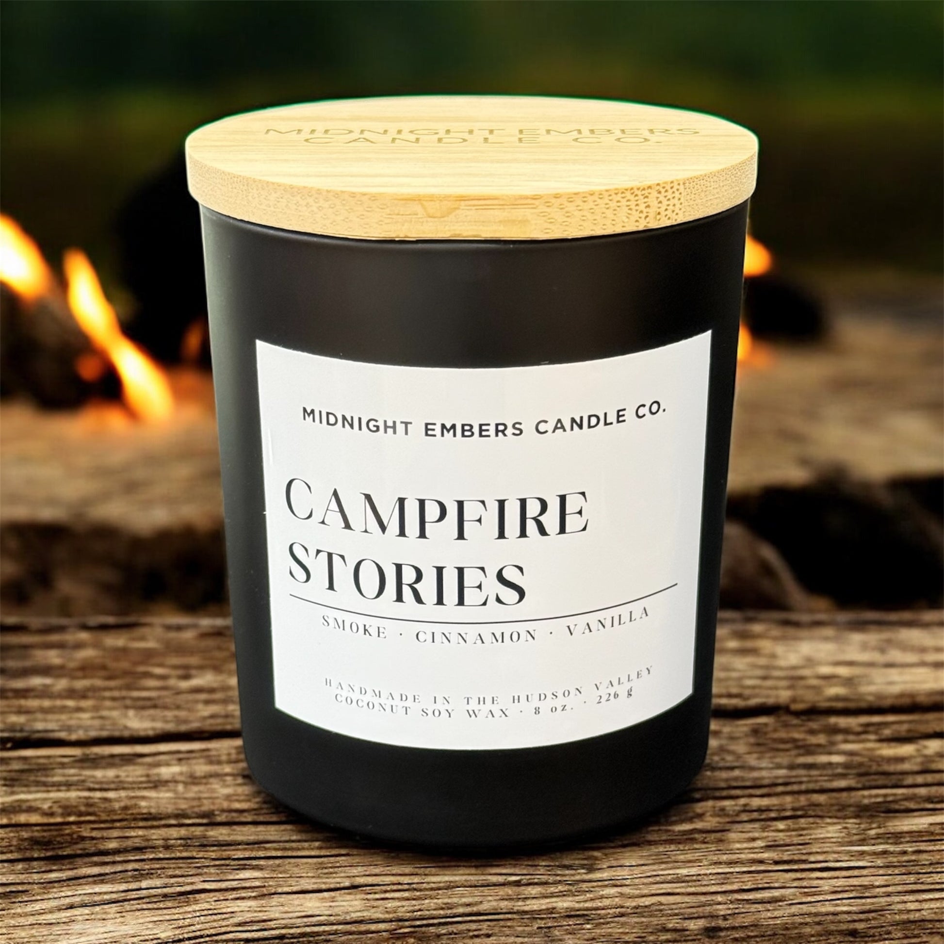 Candle labeled 'Campfire Stories' from Midnight Embers Candle Co. on a wooden surface with a campfire in the background.