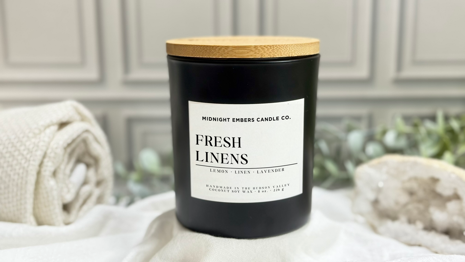 Black candle labeled 'Fresh Linens' with a wooden lid on a white surface.