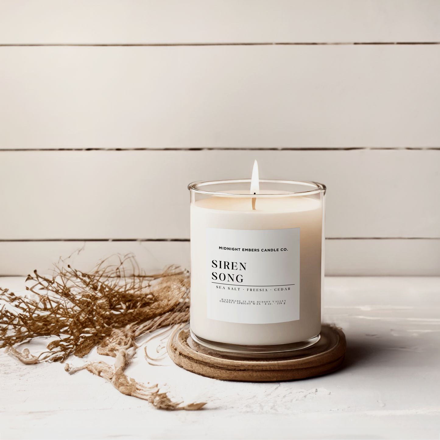 Candle labeled 'Siren Song' on a wooden coaster with dried plants on a light wooden background