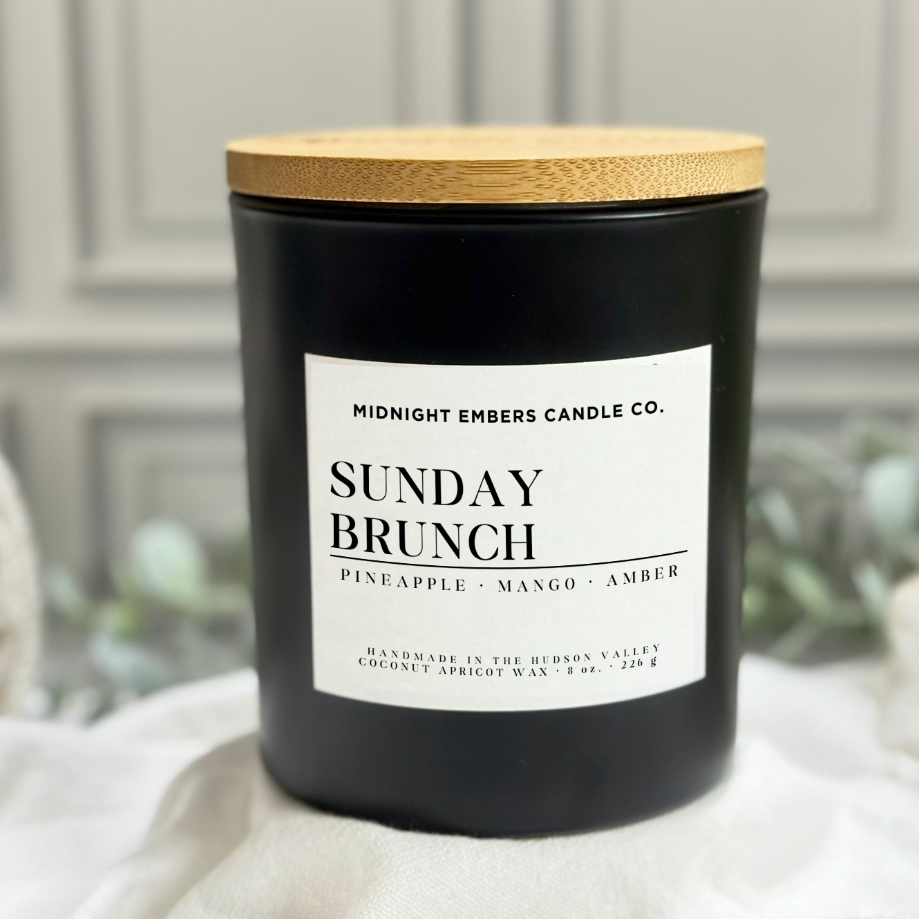 Black candle labeled 'Sunday Brunch' with a wooden lid on a white surface.