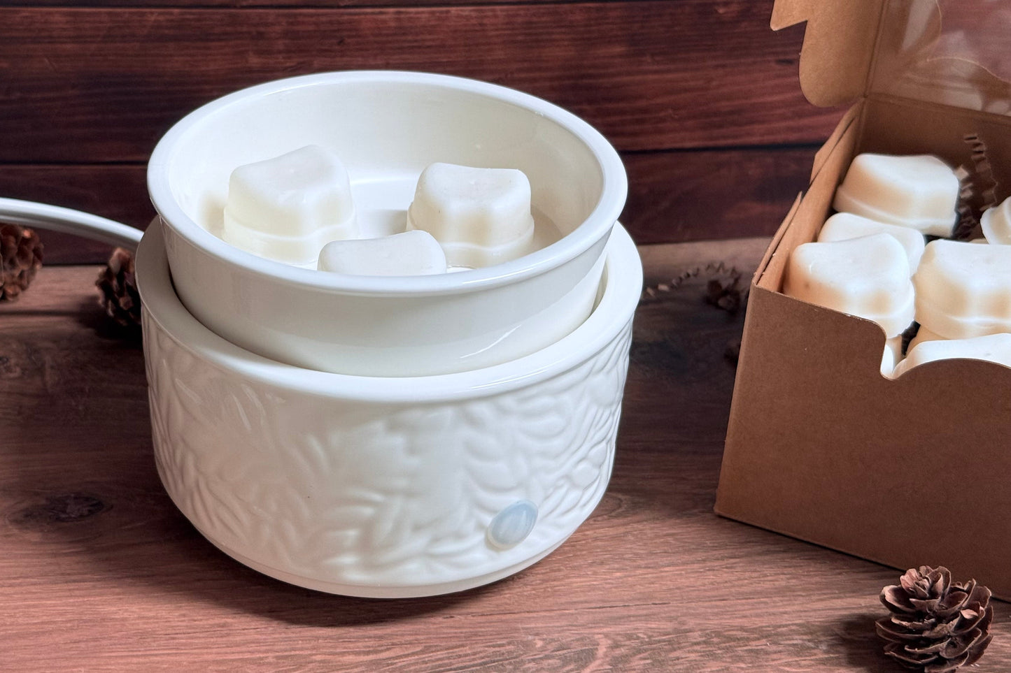 A white ceramic wax warmer with a textured base and a shallow top bowl containing wax melts, shown on a wooden surface with pinecones and a box of wax melts nearby.