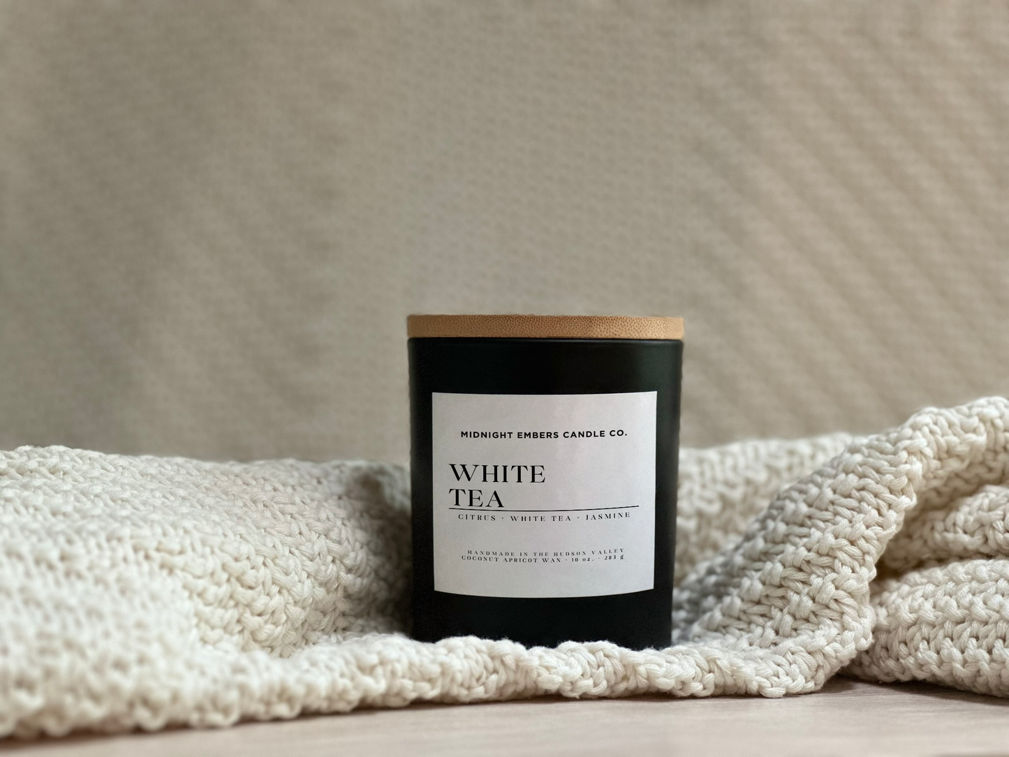 Black candle labeled 'White Tea' by Midnight Ember Candle Co. on a textured surface with a blanket.