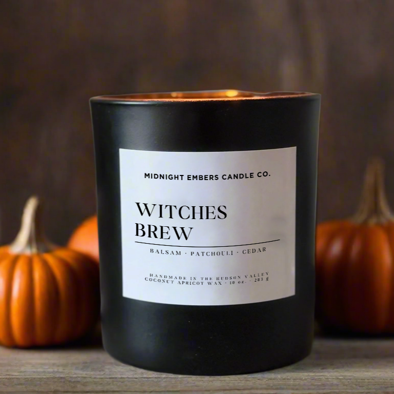 Candle labeled 'Witches Brew' by Midnight Embers Candle Co. with pumpkins and twigs in the background.